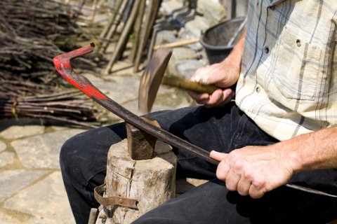 Ein Mann sitzt vor einem Holzblock und bearbeitet mit einem Hammer eine Eisenstange.