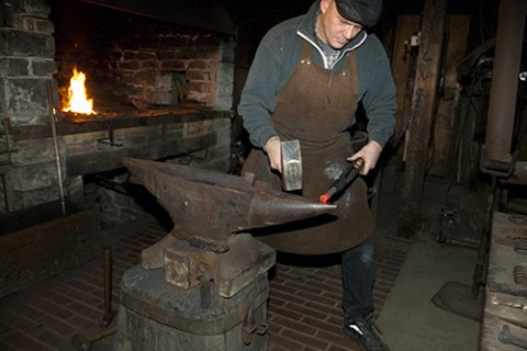 In einer Werkstatt steht ein Mann vor einem Feuer am Amboss und schmiedet glühendes Eisen. Er trägt eine Lederschürze.
