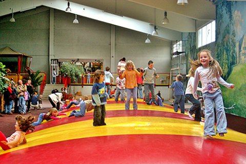 In einer Halle ist ein großes gelb-rotes Sprungkissen aufgebaut. Kinder tummeln sich darauf.
