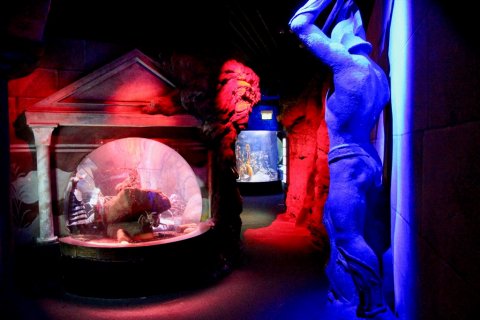Rot und blau beleuchteter, grottenartiger Gang mit einem Aquarium und einer großen Statue.
