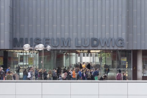 Viele Menschen stehen vor dem modernen Eingangsbereich aus Metall und Glas. In großen Buchstaben steht dort Museum Ludwig zu lesen. 