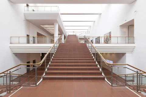 Ein Blick in das Treppenhaus des Museums. Die Treppen ist terrakottafarben gefliest, die Geländer sind aus Stahl mit einem runden Holzgriff. Wände und Emporen sind weiß gestrichen.