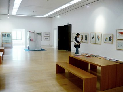 Ein Ausstellungsraum mit Exponaten, die an der Wand hängen. Im Vordergrund steht ein Tisch mit Bänken, darauf liegen ebenfalls Ausstellungsstücke. Im Hintergrund betrachtet eine Person ein Exponat an der Wand.