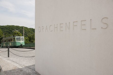 Steintafel mit der Aufschrift "Drachenfels". Im Hintergrund fährt die grüne Drachenfels-Zahnradbahn in die Bergstation ein. Noch weiter hinten sieht man den grün bewaldeten Drachenfels.
