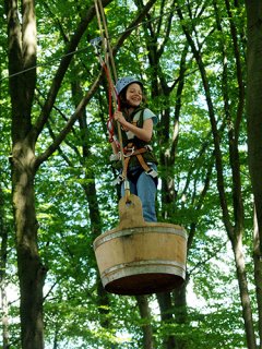 Ein Kind mit Helm schwingt in einem Holzbottich in der Luft zwischen Bäumen.