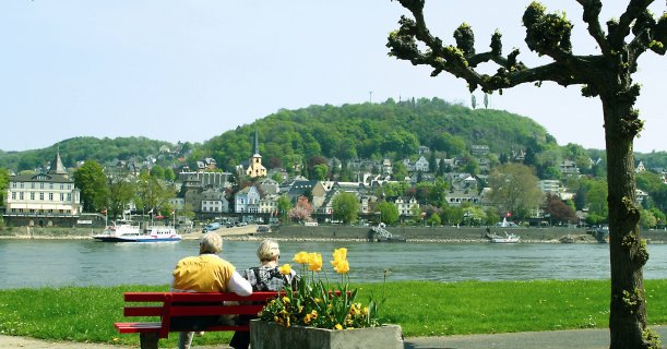 Blick von hinten auf ein älteres Paar, das linksrheinisch auf einer roten Bank sitzt, mit schöner Aussicht auf Linz. Davor ein Blumenkübel mit gelben Blumen, rechts eine Kopflinde, die zu grünen beginnt.