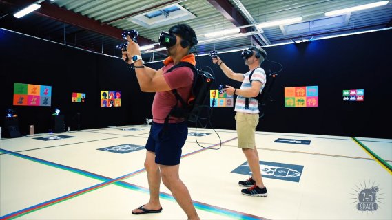 Zwei Männer stehen in einem Raum. Sie tragen VR-Brillen und zielen mit einem Spielstick auf eine Wand mit bunten Abbildungen.